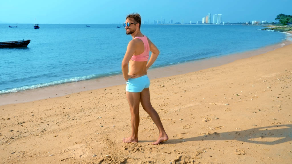 Грайливий красивий хлопець в рожевій футболці і блакитних шортах радіє на пляжі. дикий на морі
. - Фото, зображення