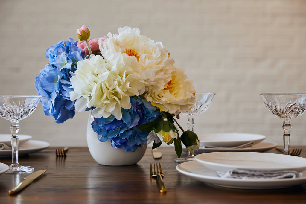 Talerze, noże i widelce, kryształowe kieliszki i bukiet w biały wazon na drewnianym stole w pobliżu Mur w domu - Zdjęcie, obraz