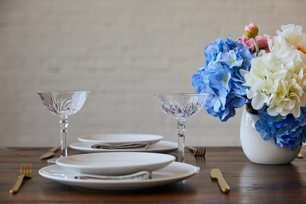 weiße Teller, Messer und Gabeln, Kristallgläser und Blumenstrauß in Vase auf Holztisch in der Nähe von Ziegelmauer zu Hause - Foto, Bild