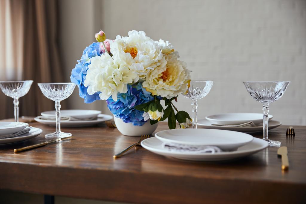platos blancos, cubiertos, vasos de cristal y ramo en jarrón sobre mesa de madera en casa
 - Foto, imagen