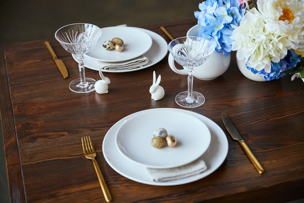 crystal glass, flowers in vase, golden cutlery and decorative rabbits on wooden table at home - Photo, Image
