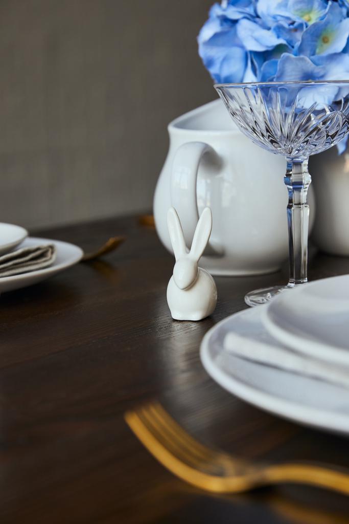 селективный фокус декоративного кролика возле тарелок и хрусталя на деревянном столе в домашних условиях
 - Фото, изображение