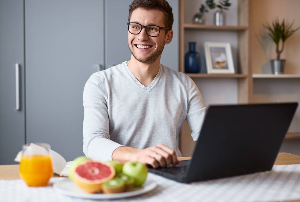 Усміхнений хлопець переглядає ноутбук під час сніданку
 - Фото, зображення
