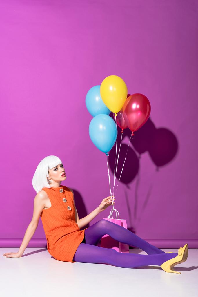 Стильная молодая женщина в парике держит воздушные шары и сидит на фиолетовом фоне
 - Фото, изображение