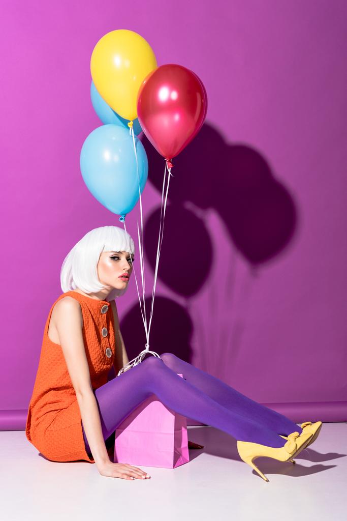 Chica seria en peluca blanca con globos de aire sentado sobre fondo púrpura
 - Foto, imagen