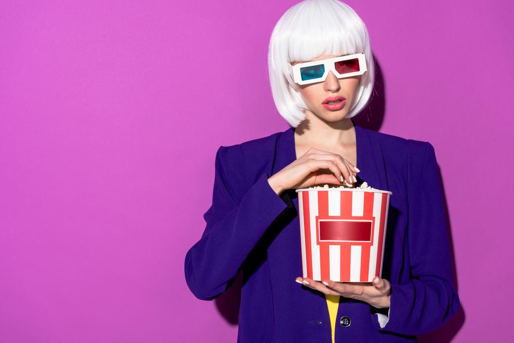 Прекрасная молодая женщина в 3D-очках ест попкорн на фиолетовом фоне
 - Фото, изображение