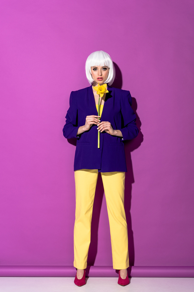 Серьёзная девушка в синей куртке держит желтый цветок на фиолетовом фоне
 - Фото, изображение