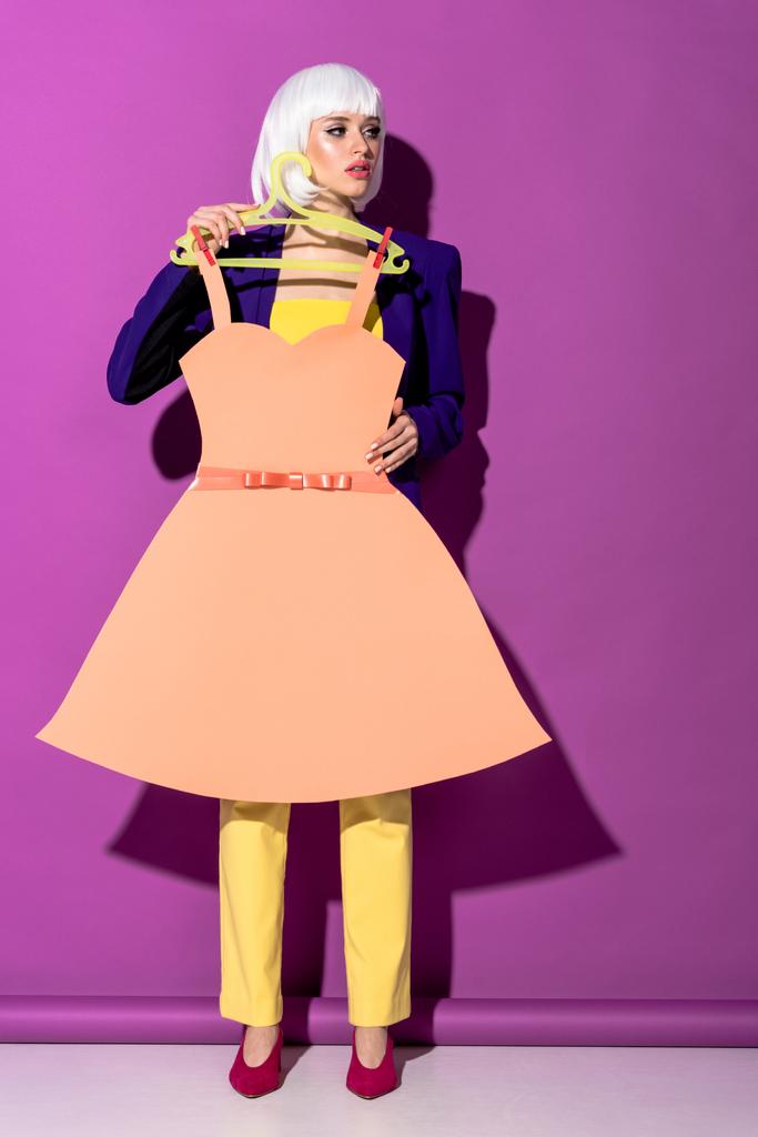 Prachtige jonge vrouw in witte pruik bedrijf hanger met papieren jurk op paarse achtergrond - Foto, afbeelding