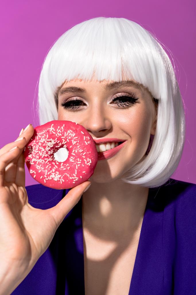 Lachende vrouw in pruik houden van roze donut geïsoleerd op paars - Foto, afbeelding