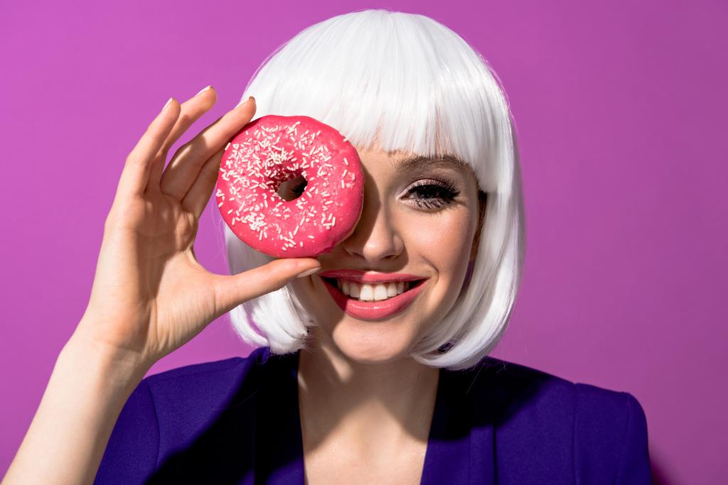 Mooi meisje roze donut houden op paarse achtergrond glimlachen - Foto, afbeelding