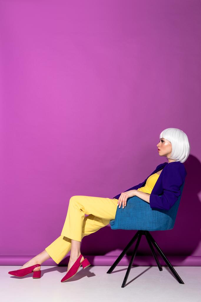 Vista lateral de chica con estilo en peluca blanca sentado en sillón sobre fondo púrpura
 - Foto, imagen