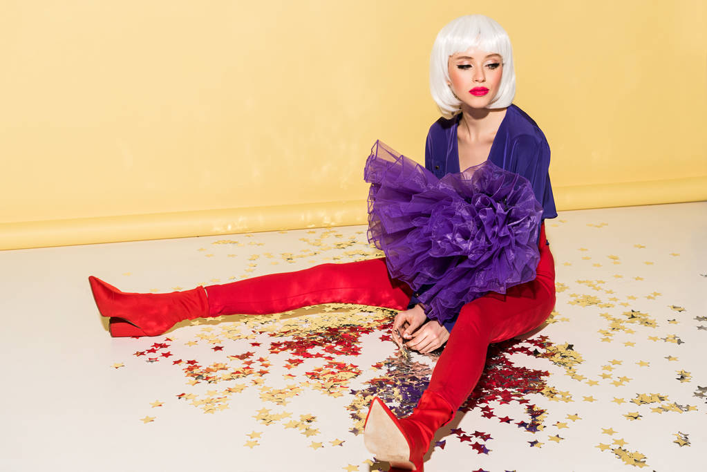 Pensive woman in wig and red pants sitting on star confetti on yellow background - Photo, Image