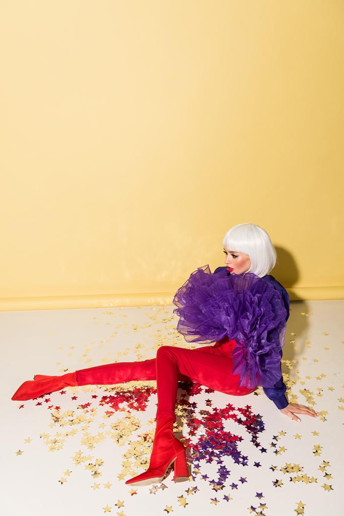 Young woman in red pants sitting on sparkle confetti on yellow background - Photo, Image