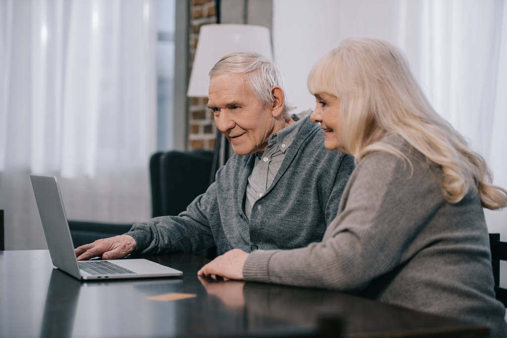 счастливая пожилая пара, сидящая за столом и с ноутбуком дома
 - Фото, изображение