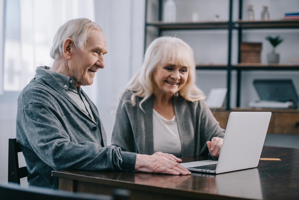sonriente pareja de ancianos sentado en la mesa y el uso de ordenador portátil en casa
 - Foto, Imagen