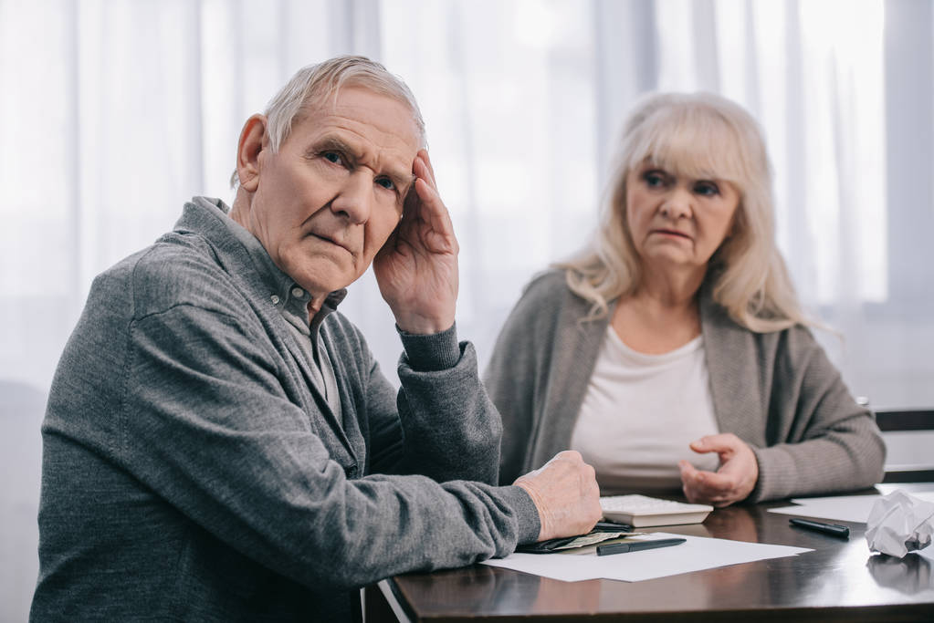 Senior man met de hand op het hoofd zitten aan tafel in de buurt van facturen met vrouw op achtergrond - Foto, afbeelding