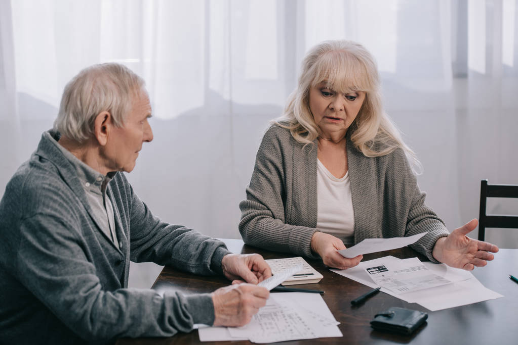 surprised senior couple in casual clothes sitting at table and holding bills at home - Photo, Image