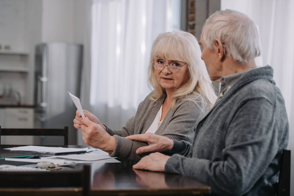 mujer mayor en gafas sosteniendo facturas y mirando a la cámara mientras está sentado en la mesa con el hombre
 - Foto, Imagen