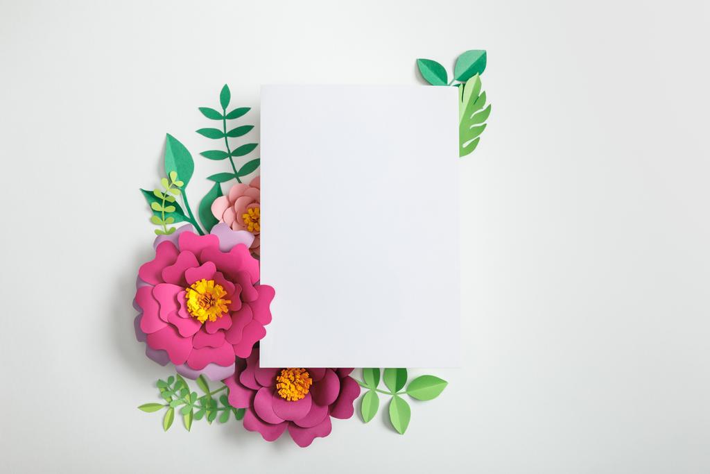 top view of white blank card near pink paper flowers and green leaves on grey background - Photo, Image