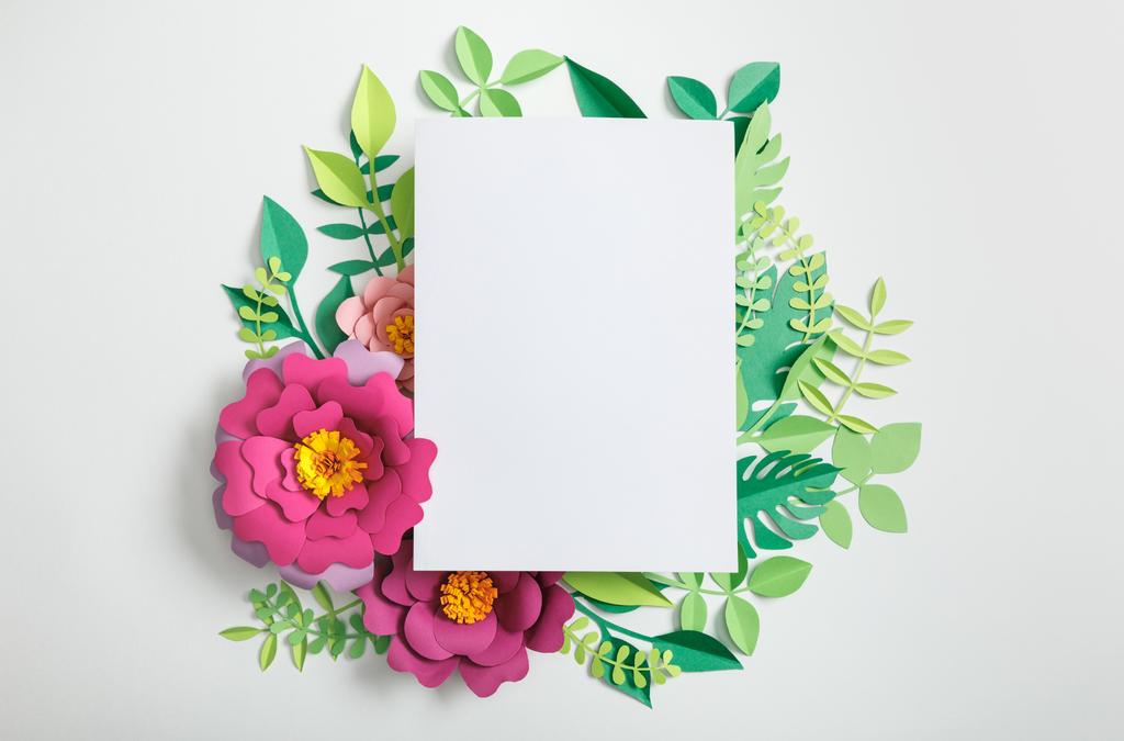 top view of white blank card on paper flowers and green leaves on grey background - Photo, Image