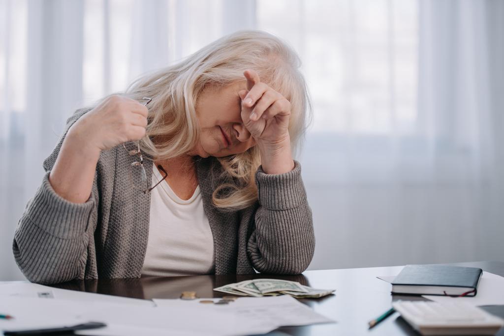 втомилася старша жінка сидить за столом і має головний біль під час підрахунку грошей вдома
 - Фото, зображення