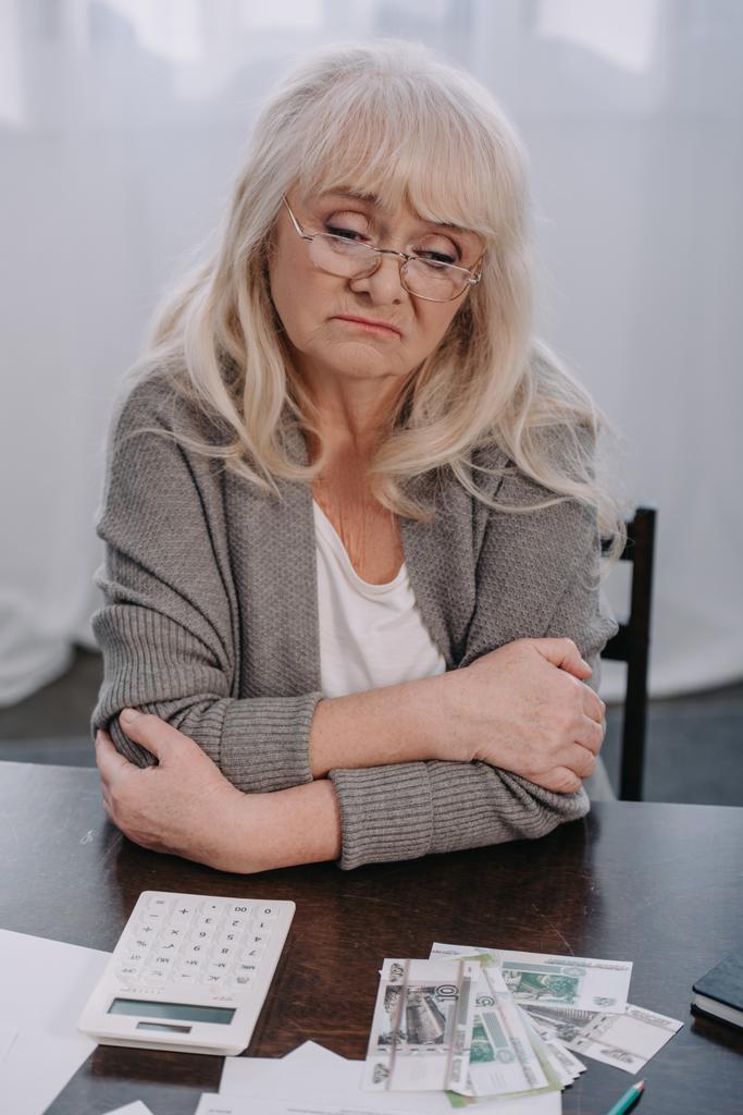 donna anziana sconvolta con le braccia incrociate seduta a tavola con soldi e calcolatrice
  - Foto, immagini