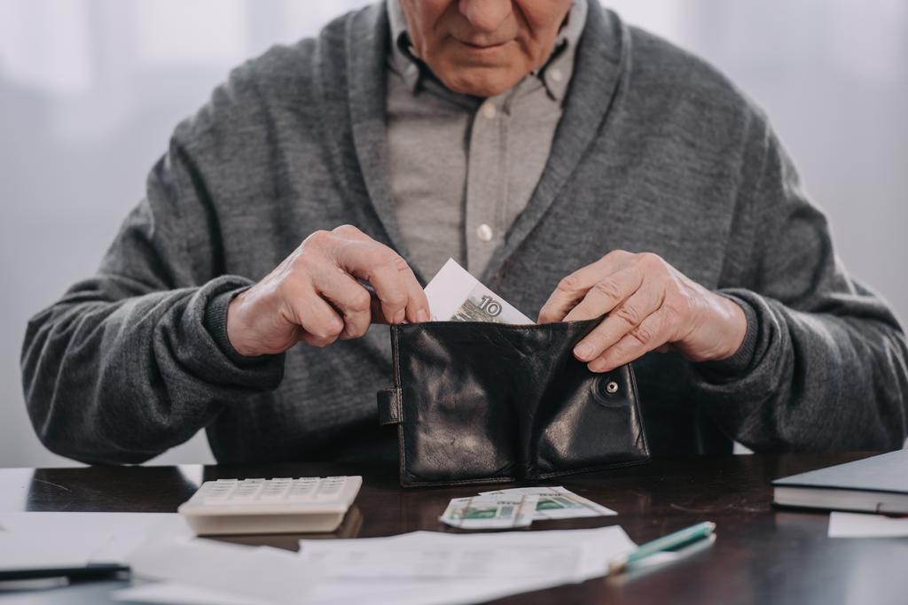 bijgesneden weergave van mannelijke gepensioneerde zittend aan tafel met papierwerk en brengen geld in de portemonnee  - Foto, afbeelding