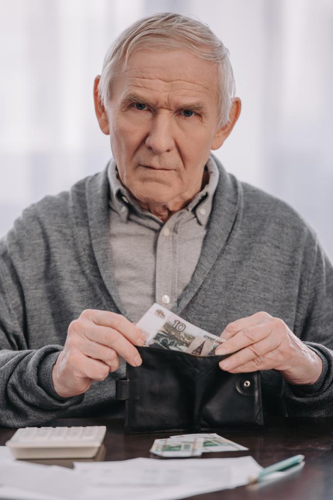pensionista masculino sentado en la mesa, mirando a la cámara y poniendo dinero en la cartera
  - Foto, imagen