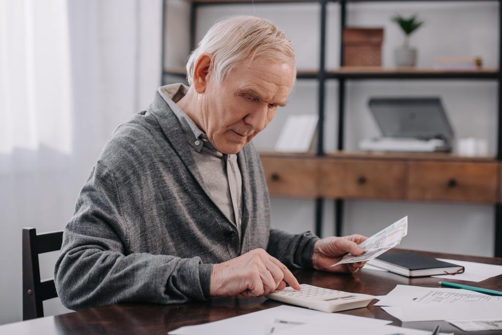 idősebb ember ül az asztal, a papírmunka és a számológép használata közben számolja a pénzt  - Fotó, kép