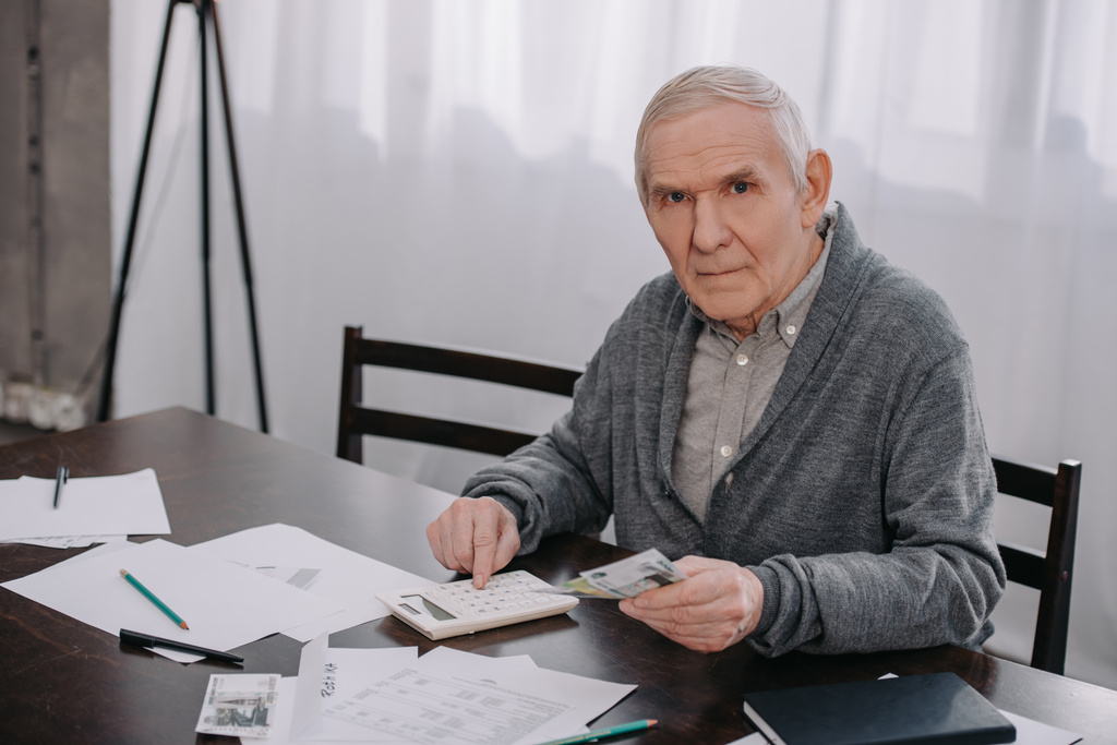 старший чоловік у повсякденному одязі, сидячи за столом з паперовою роботою та використовуючи калькулятор під час підрахунку грошей
  - Фото, зображення