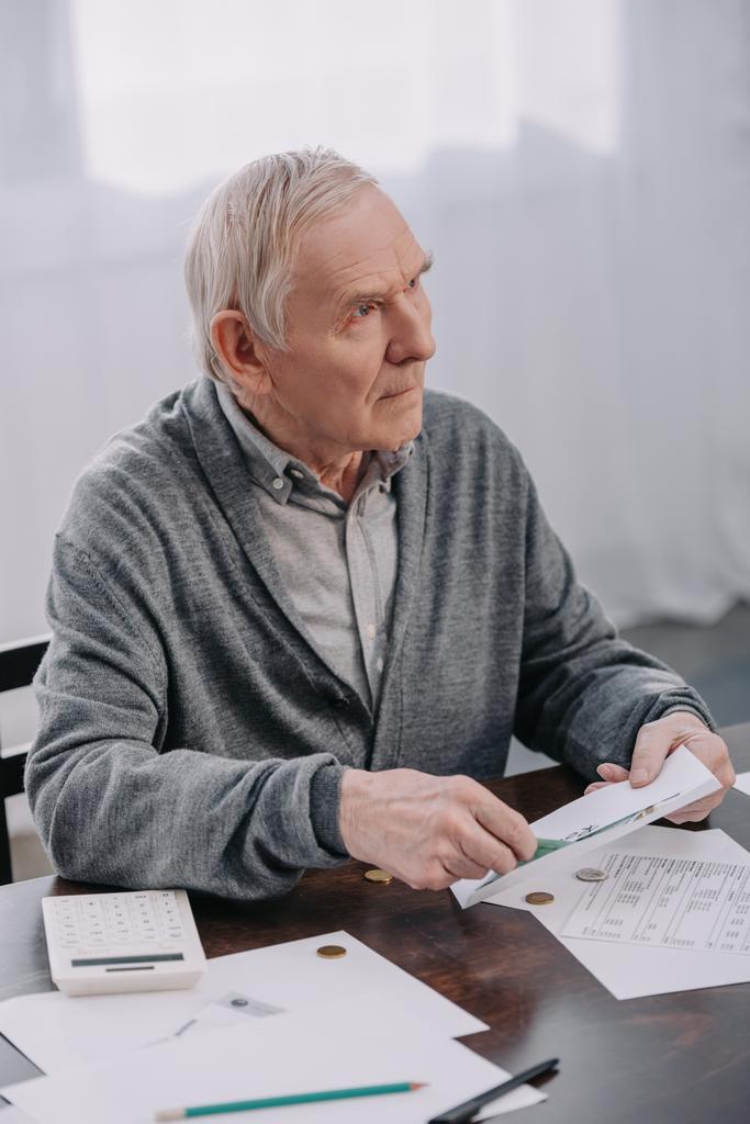 Senior sitzt mit Papierkram am Tisch und hält Umschlag beim Geldzählen in der Hand - Foto, Bild