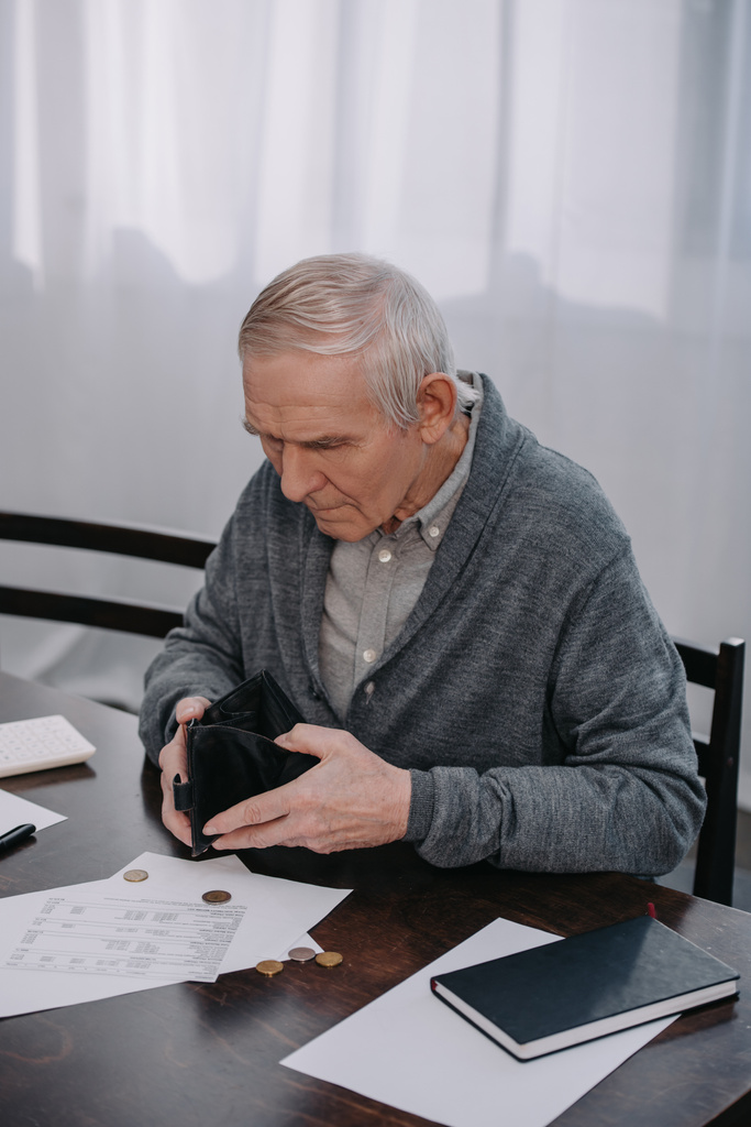 Rentnerin sitzt mit Papierkram am Tisch und hält leere Brieftasche  - Foto, Bild