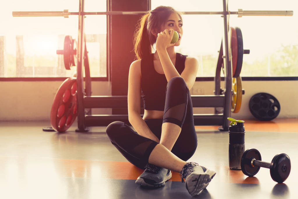 Жінка тренується в фітнес-тренажерному залі, розслабляється, тримаючи яблучні фрукти після тренувальних видів спорту з гантелі та білковою пляшкою для струшування здорового способу життя
. - Фото, зображення