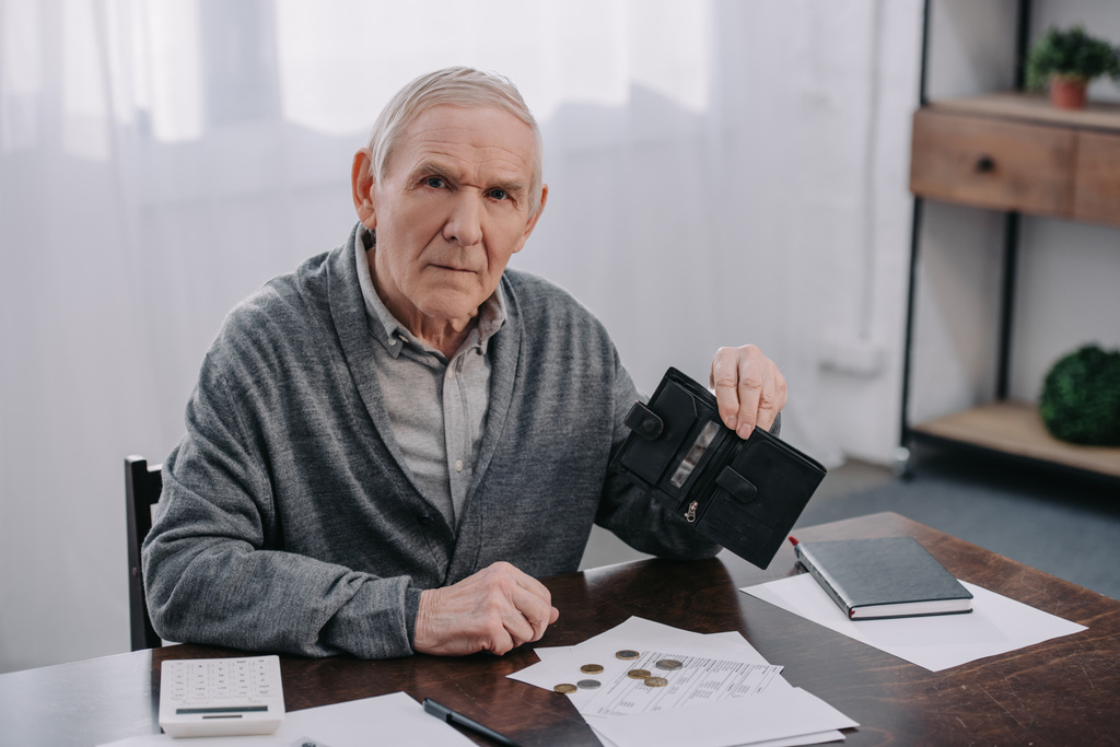 старший чоловік сидить за столом з папером і тримає гаманець, рахуючи гроші вдома
 - Фото, зображення