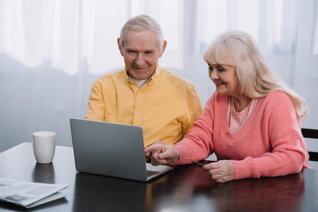 sonriente pareja de ancianos en ropa casual usando el ordenador portátil mientras está sentado en la mesa en casa
 - Foto, imagen