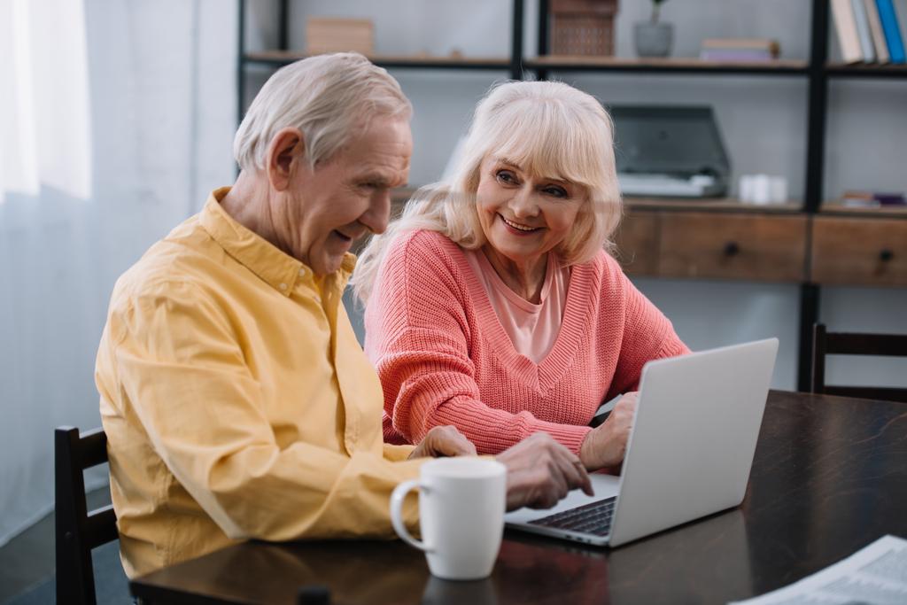 glimlachend senior paar in casual kleding met behulp van laptop zittend aan tafel thuis - Foto, afbeelding