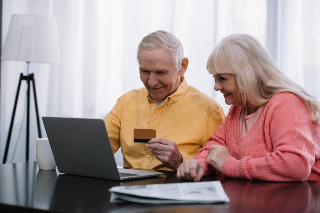 pareja de ancianos sentados en el sofá con el ordenador portátil y la tarjeta de crédito, mientras que hacer compras en línea en casa
 - Foto, Imagen