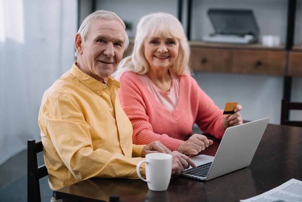 старша пара сидить на дивані з ноутбуком і кредитною карткою, дивлячись на камеру і роблячи онлайн покупки вдома
 - Фото, зображення