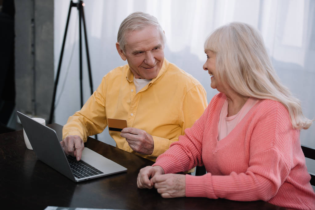 щаслива старша пара сидить на дивані з ноутбуком і кредитною карткою під час онлайн-покупки вдома
 - Фото, зображення