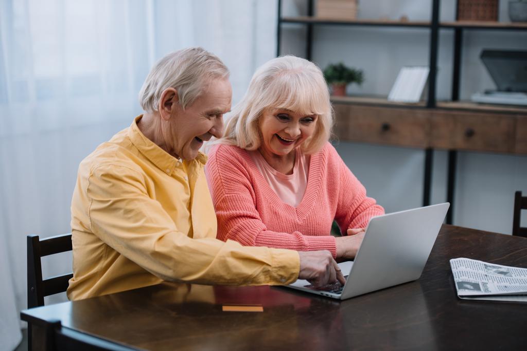 hymyilevä vanhempi pari käyttää kannettavaa tietokonetta istuessaan pöydässä kotona
 - Valokuva, kuva