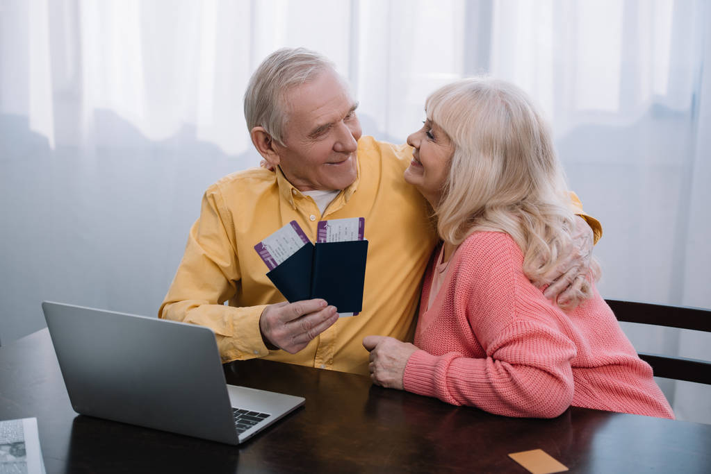 自宅のパスポートと航空券をハグとノート パソコンとテーブルに座っている老夫婦の笑顔  - 写真・画像