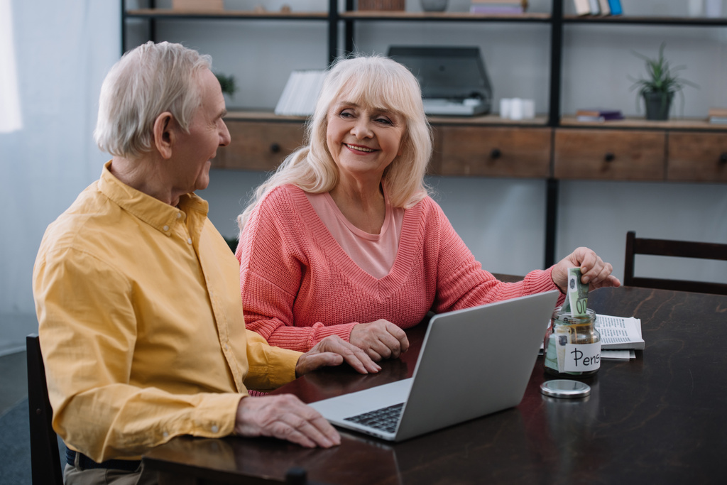 idősebb pár ül asztal, laptop, miközben pénzt öltünk bele a "nyugdíj" betűkkel üvegedénybe nő - Fotó, kép