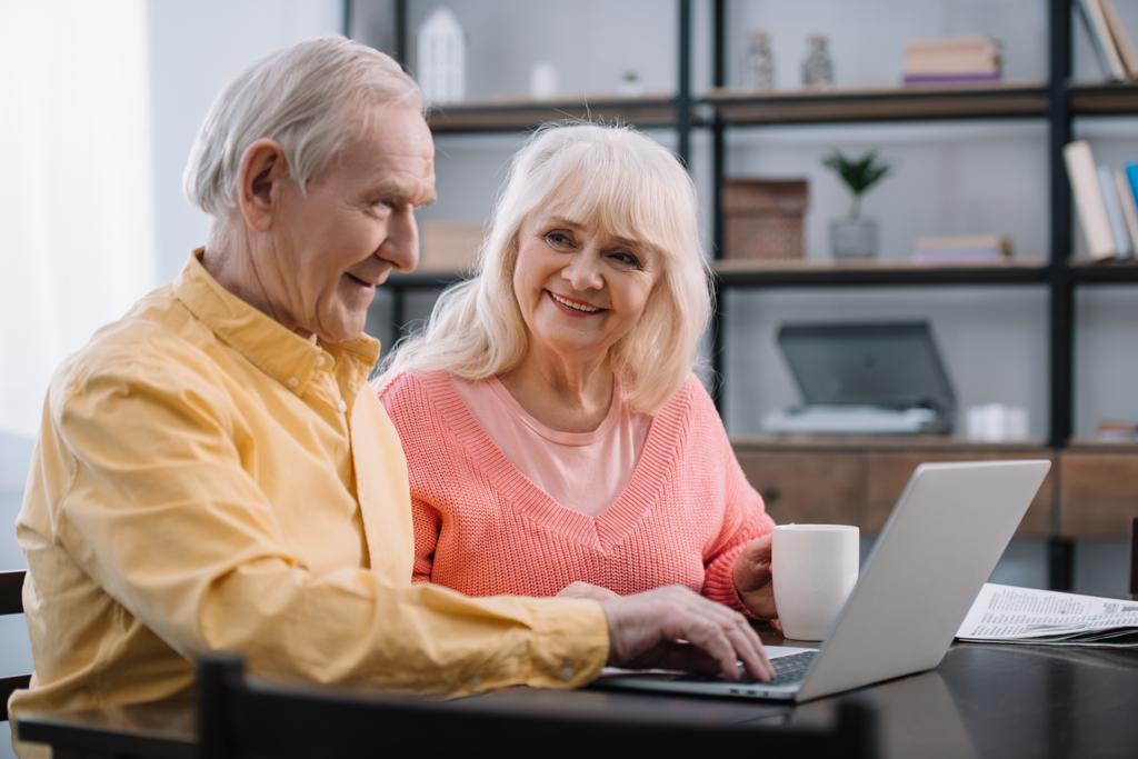 улыбающаяся пожилая пара в красочной одежде, сидящая за столом и пользующаяся ноутбуком
 - Фото, изображение