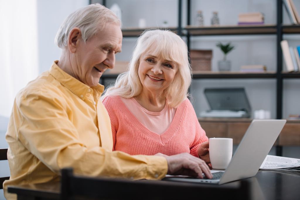 happy senior couple in colorful clothes sitting at table and using laptop - Photo, Image
