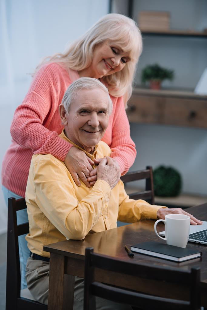 Senior vrouw knuffelen Glimlachende man aan tafel zitten en kijken naar camera - Foto, afbeelding