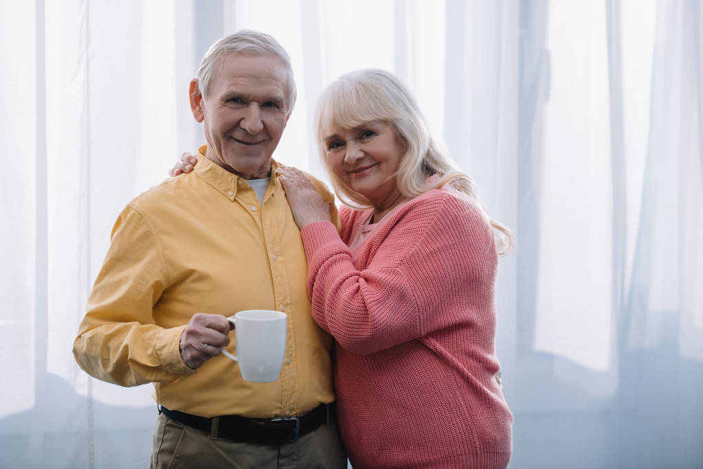 usměvavá starší žena s šedými vlasy, při pohledu na fotoaparát a všeobjímající muž s šálkem čaje doma - Fotografie, Obrázek