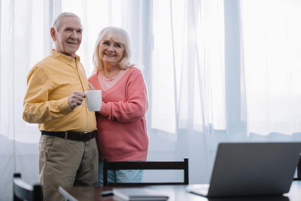 Selektiver Fokus eines lächelnden Seniorehepaares in lässiger Kleidung, das zu Hause eine Tasse Kaffee mit Kopierplatz hält - Foto, Bild