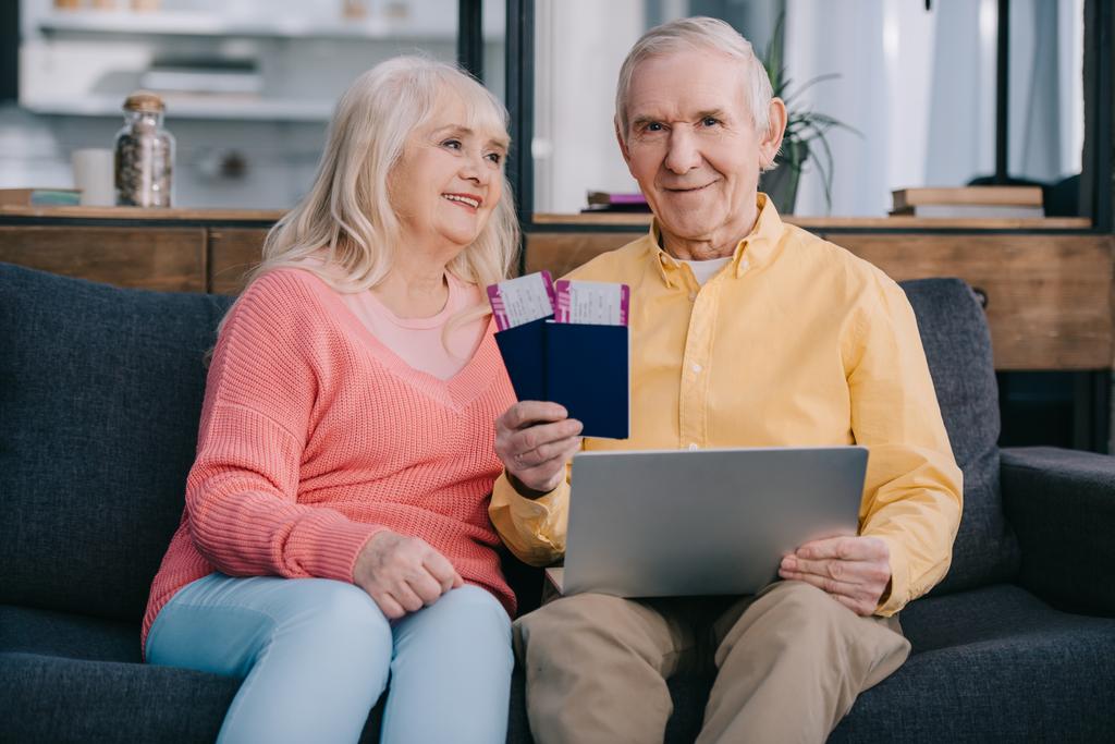 улыбающаяся пожилая пара с авиабилетами, ноутбуками и паспортами, сидя дома на диване
  - Фото, изображение