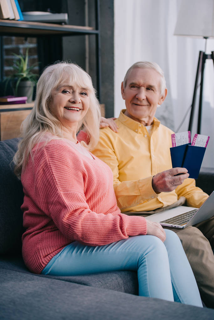улыбающаяся пожилая пара сидит на диване с авиабилетами и паспортами дома
  - Фото, изображение