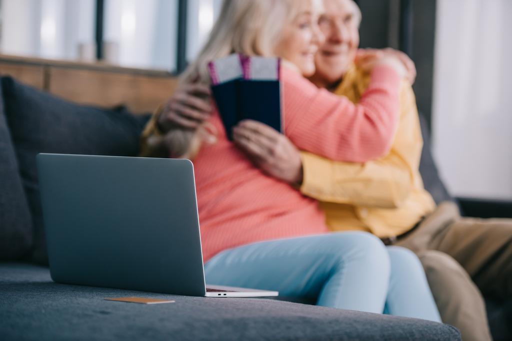 selektiver Fokus von Laptop und Kreditkarte mit einem älteren Paar, das sich umarmt, während es Flugtickets und Pässe im Hintergrund hält - Foto, Bild